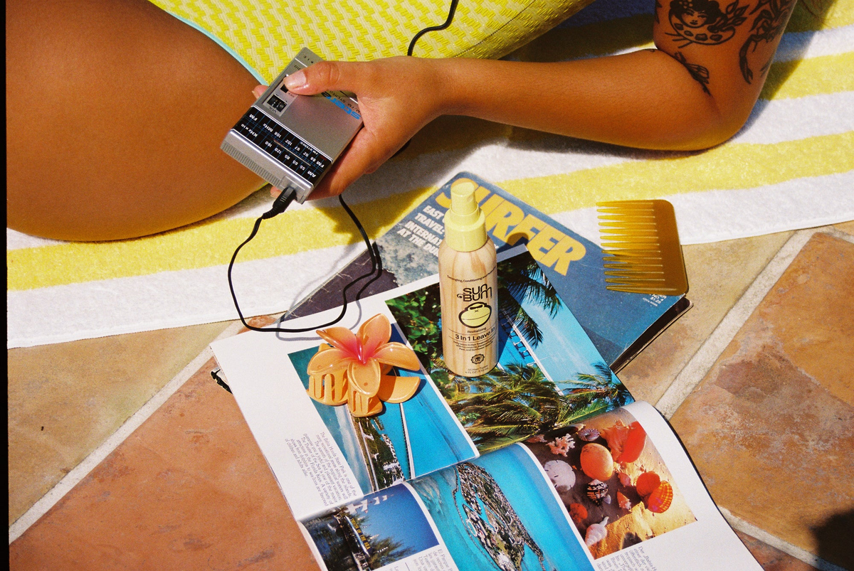 A person lying on a yellow and white striped towel on a tiled floor, holding a silver cassette tape